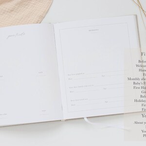May include: An open baby memory book with white pages featuring printed lines and headings for recording milestones. The left page is titled "your firsts" and the right page includes a section for "our story" with prompts for dates and ages.
