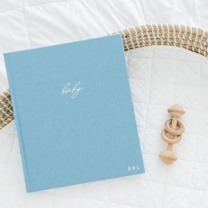 May include: A light blue hardcover book with the word baby written in white cursive script on the front cover and the letters BBL printed in white in the bottom right corner, accompanied by a small wooden ring rattle toy with three rings.