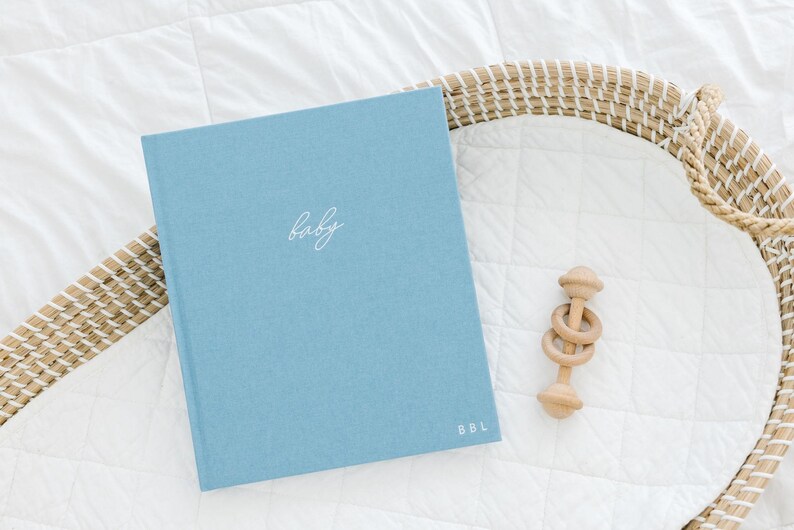 May include: A light blue hardcover book with the word baby written in white cursive script on the front cover and the letters BBL printed in white in the bottom right corner, accompanied by a small wooden ring rattle toy with three rings.