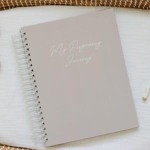May include: A light taupe spiral-bound journal with the text My Pregnancy Journey printed in white script on the front cover, accompanied by a beige plush crescent moon toy and a slim white pen with gold accents placed on a white fabric surface.