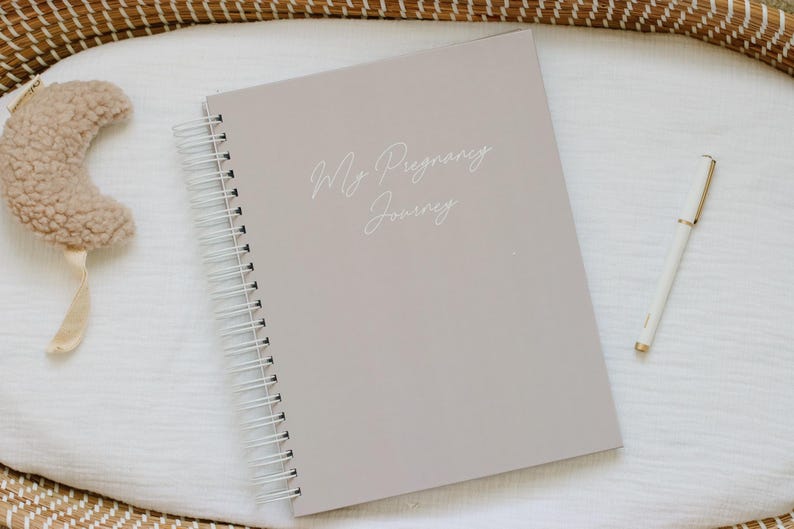 May include: A light taupe spiral-bound journal with the text My Pregnancy Journey printed in white script on the front cover, accompanied by a beige plush crescent moon toy and a slim white pen with gold accents placed on a white fabric surface.