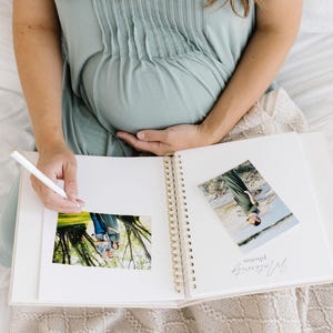 May include: A white spiral-bound photo album with gold-colored rings, featuring two photographs attached to the pages. One page displays the text "Maternity photos" in a cursive script. A white pen is held over the open album pages.
