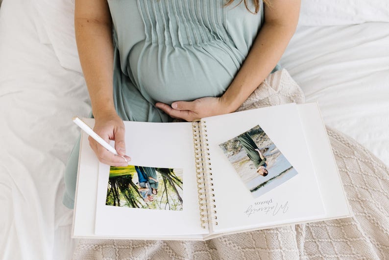 May include: A white spiral-bound photo album with gold-colored rings, featuring two photographs attached to the pages. One page displays the text "Maternity photos" in a cursive script. A white pen is held over the open album pages.
