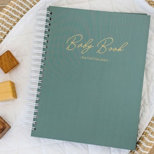 May include: A teal green baby book with gold lettering that reads "Baby Book - the first five years -" on the cover. The book is lying on a white surface with a woven basket and wooden blocks.