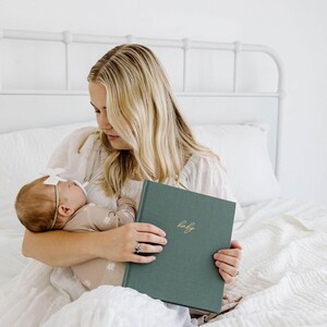 May include: A rectangular, forest green fabric-covered baby memory book with the word baby embossed in gold cursive script on the front cover. The book has a hardbound spine and a textured, cloth-like finish, held in a neutral-toned setting.