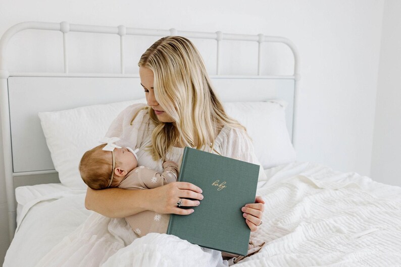 May include: A rectangular, forest green fabric-covered baby memory book with the word baby embossed in gold cursive script on the front cover. The book has a hardbound spine and a textured, cloth-like finish, held in a neutral-toned setting.