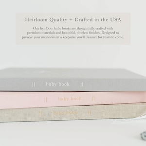 May include: Three rectangular baby books stacked horizontally, featuring gray, light pink, and beige covers. Each spine displays the text "baby book" with vertical lines. Above the stack, text reads "Heirloom Quality + Crafted in the USA."