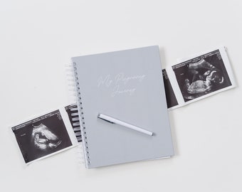 May include: A gray spiral bound journal with the text "My Pregnancy Journey" on the cover. Two black and white ultrasound images are on a white surface next to the journal.