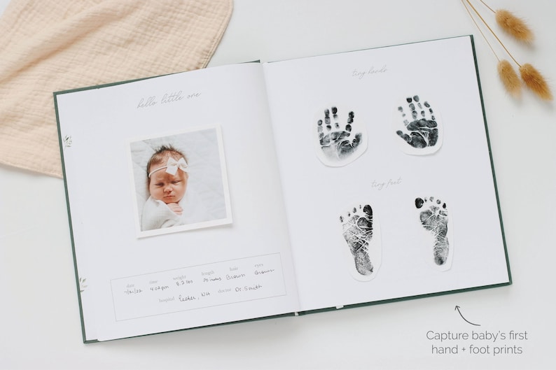 May include: An open baby memory book with a dark green cover. The left page features a square photograph of a baby with a white bow, and a printed box for birth details. The right page displays two black ink handprints and two footprints.