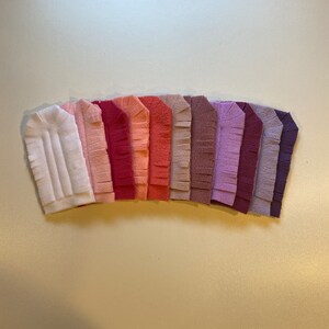 May include: A set of ten felt fabric strips in various shades of pink, purple, and white. Each strip has a fringed edge and is arranged in a gradient pattern.