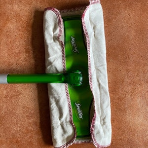 Op de afbeelding: Een witte Swiffer Sweeper mopkop met een groene kunststofsteel en het woord "Swiffer" gedrukt op de steel. De mopkop is met rode stiksels aan de steel bevestigd.