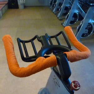 Op de afbeelding: Oranje gevulde handvatten op een stationaire trainingsfiets met een zwart frame en een waterfleshouder.