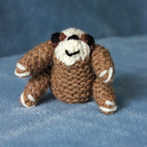 May include: A small crocheted sloth toy with brown and white fur. The sloth has a smiling face with black eyes and a brown nose.