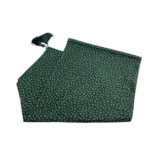 Puede incluir: Un camino de mesa verde oscuro con un patrón repetido de bastones de caramelo y hojas de acebo. El camino de mesa tiene una borla verde oscuro en un extremo. Ideal para la temporada navideña.