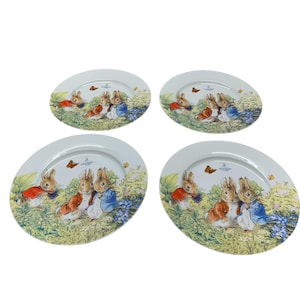 May include: Four white ceramic plates with a Peter Rabbit design. Each plate features illustrations of rabbits in a garden setting with butterflies and flowers. The plates have a slightly oval shape.