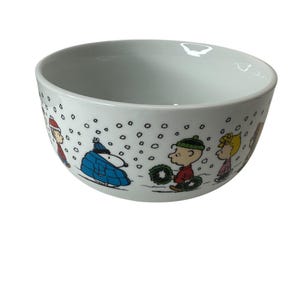 Puede incluir: Cuenco de cer&aacute;mica blanca con interior gris claro, decorado con personajes de Peanuts en una escena invernal. El cuenco presenta ilustraciones de Snoopy, Charlie Brown y otros personajes, con copos de nieve cayendo.