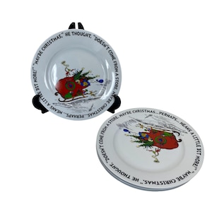 May include: White ceramic plates with a Dr. Seuss-inspired design. The plates feature a red sleigh with a Christmas tree and the quote, "Maybe Christmas...perhaps...means a little bit more!" Black plate stands hold the plates.