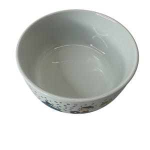 Puede incluir: Cuenco de cer&aacute;mica blanco con forma redondeada. El cuenco tiene un interior liso y brillante y una banda decorativa alrededor del exterior con figuras de dibujos animados y detalles en azul y negro. El cuenco est&aacute; vac&iacute;o.