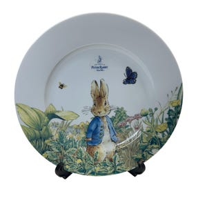 May include: White ceramic plate featuring Peter Rabbit in a blue jacket, surrounded by a garden scene with a bee and butterfly. The plate has the text "The World of Peter Rabbit" in blue.