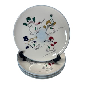 Puede incluir: Juego de cuatro platos de cerámica blanca con borde azul claro. Cada plato presenta una ilustración caprichosa de muñecos de nieve patinando sobre hielo, con bufandas, sombreros y mitones coloridos. Un encantador juego de vajilla con temática invernal.