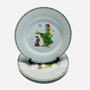 May include: Set of white ceramic plates featuring a colorful dotted border. Each plate has a central illustration of a green character with a Christmas tree and a dog wearing Santa hats. Festive holiday dinnerware.