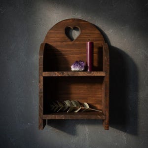 May include: A dark stained wooden shelf with a heart-shaped cutout. A purple candle and amethyst crystal sit on the top shelf. A sprig of greenery rests on the bottom shelf. The shelf is mounted on a dark gray wall.