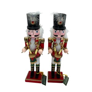 May include: Two wooden nutcracker figurines with black hats, white beards, and red and gold uniforms. They are holding drumsticks and drums. The nutcrackers are standing on red bases with gold trim.