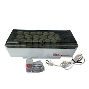 Op de afbeelding: Een witte en zwarte Deluxe Express Hairsetter met een doorzichtig deksel, met meerdere haarrollers. Het apparaat heeft een netsnoer en een klein zakje met accessoires. De tekst "Deluxe Express Hairsetter" staat op de zijkant.