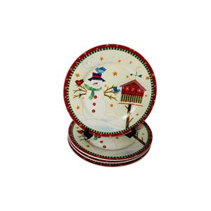 May include: A stack of round ceramic plates featuring a festive winter scene with a central snowman wearing a blue hat and green scarf, a red birdhouse labeled Merry Christmas, small blue birds, yellow stars, and a red and green striped border.