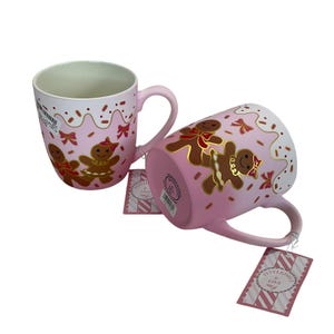 Puede incluir: Dos tazas de cerámica rosa con diseños de hombre de jengibre. Una taza está en posición vertical, la otra está de lado. Las tazas tienen un fondo rosa con glaseado blanco y chispas. Los hombres de jengibre están decorados con detalles dorados y lazos rojos.