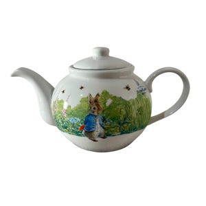Può includere: Teiera in ceramica bianca con un design di Peter Rabbit. La teiera ha un beccuccio e un manico curvi, un coperchio e una scena dipinta di Peter Rabbit in un giardino con api. Un design classico per servire il tè.