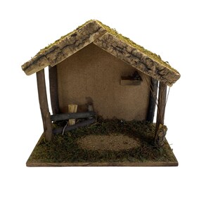 Op de afbeelding: Een miniatuur houten bouwwerk met een zadeldak bedekt met groen mos, een getextureerde bruine achterwand en een basis bedekt met mos. Het open gebouw bevat een kleine houten plank, een hek en een bundel stro.