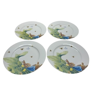 May include: Four white ceramic plates with a Peter Rabbit design. Each plate features an illustration of Peter Rabbit in a blue jacket, surrounded by green leaves, yellow flowers, and bees. The plates are round with a slightly raised edge.