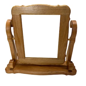 May include: A wooden, rectangular picture frame with a light brown finish. The frame has a decorative, scalloped top and is supported by two curved side supports and a base. The frame is designed to rotate.