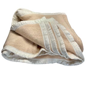 May include: A folded beige blanket with a white satin trim. The blanket is made of a soft, fuzzy material and has a quilted border. The blanket is folded over on itself, showing the texture and design.