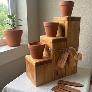 May include: A tiered wooden plant stand with three terracotta pots of varying sizes. The stand has a rustic design, with a twine accent and wooden tags that read "Happy Mother's Day" and "Handmade Wisconsin". Three wooden plant markers are also present.