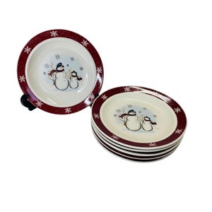 May include: Set of six white ceramic plates with a burgundy rim and snowflake design. Each plate features a snowman family illustration in the center. The plates are stacked and displayed on a white background.