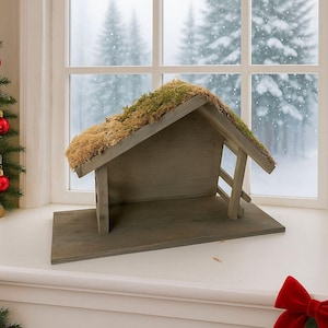 May include: A wooden nativity stable with a moss-covered roof. The grey structure has a simple design with a sloped roof and open front. The background shows a snowy winter scene through a window. The stable is on a windowsill.