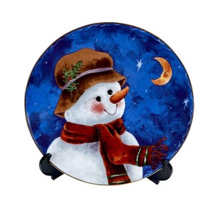 May include: A round decorative plate featuring a snowman with a brown hat, orange carrot nose, and red scarf. The background is a dark blue night sky with a crescent moon and stars. The plate is displayed on a black stand.