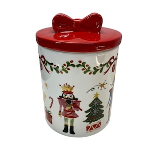 Può includere: Un barattolo natalizio in ceramica bianca con coperchio rosso e manico a forma di fiocco. Il barattolo è decorato con illustrazioni festive di uno schiaccianoci, un albero di Natale e altri elementi natalizi. Il barattolo è di forma cilindrica.