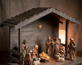 Könnte beinhalten: Ein hölzerner Krippenstall mit Giebeldach und einem Sternsymbol an der Rückwand. Die Struktur besteht aus einer Bodenplatte und Seitenstützen. Der Text unten rechts lautet: Figurines aren't included — stable only. Das Holz hat ein dunkles Finish.