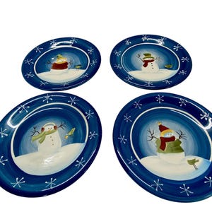 May include: Set of four oval ceramic plates with a dark blue border and a white interior. Each plate features a snowman design with a hat, scarf, and bird. White snowflakes are painted around the border.