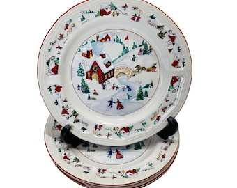 May include: A stack of white ceramic plates with a red rim and a winter scene design. The plates feature a village scene with a church, bridge, and people ice skating. The plates are on a black stand.