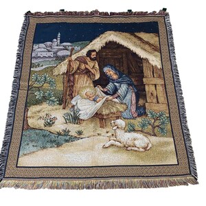 Pode incluir: Cobertor de tapeçaria tecida retratando a cena da Natividade. O cobertor apresenta uma ilustração detalhada de Maria, José e o menino Jesus em uma manjedoura, com um cordeiro e uma vila ao fundo. O cobertor tem uma borda azul escura e franjas.
