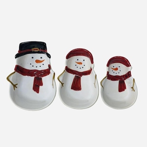 May include: Set of three snowman-shaped serving dishes. Each snowman has a white body, a red scarf, and an orange carrot nose. The largest snowman wears a black hat, while the others wear red hats. The dishes have gold-colored arms.