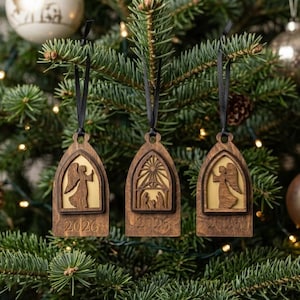 May include: Three dark brown wooden ornaments with black ribbon hangers. Each features an arched cutout design with a light-coloured backing. Two ornaments show an angel silhouette, and one shows a nativity scene. The year 2026 is engraved on each.