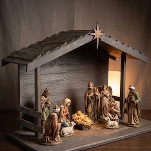 May include: A wooden nativity scene set featuring a stable structure with a star at the peak. The set includes hand-painted figurines of the holy family, three wise men, an angel, and a lamb, all arranged on a rectangular wooden base.