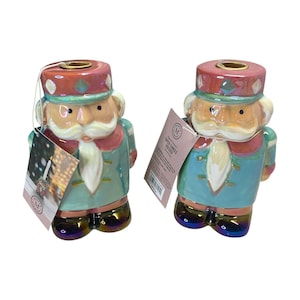 May include: A pair of ceramic nutcracker candle holders. The holders are painted in shades of pink, blue, and green with gold accents. The nutcrackers are wearing blue and red uniforms with white trim. The candle holders are labeled "Tea Light Candle Holder".