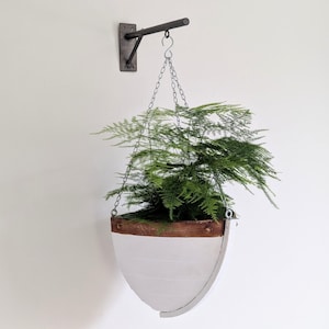 Op de afbeelding: Een hangende plantenbak met een wit, bootvormig ontwerp en een bruine houten rand. De plantenbak wordt opgehangen door een zilveren ketting aan een zwarte metalen beugel die aan de muur is bevestigd. De plantenbak is gevuld met groene, bladrijke planten.