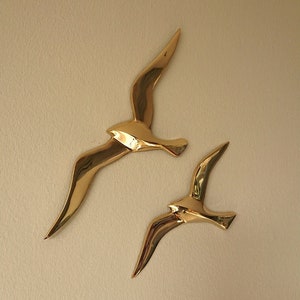 May include: Two gold metal wall sculptures of birds in flight. The birds are stylized and abstract, with long, curved wings.